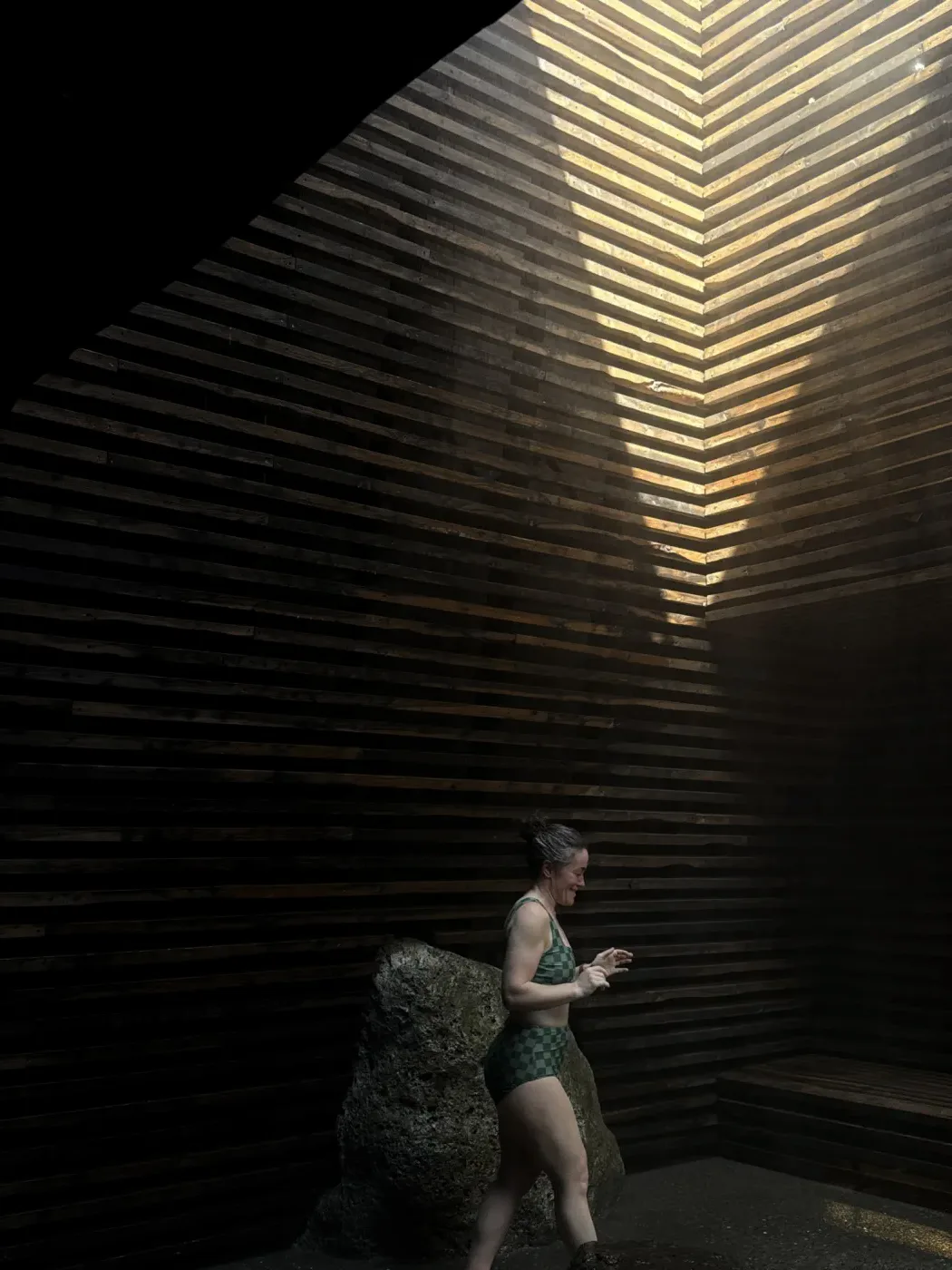A woman stepping out of a dark timber sauna with light slanting through the slats