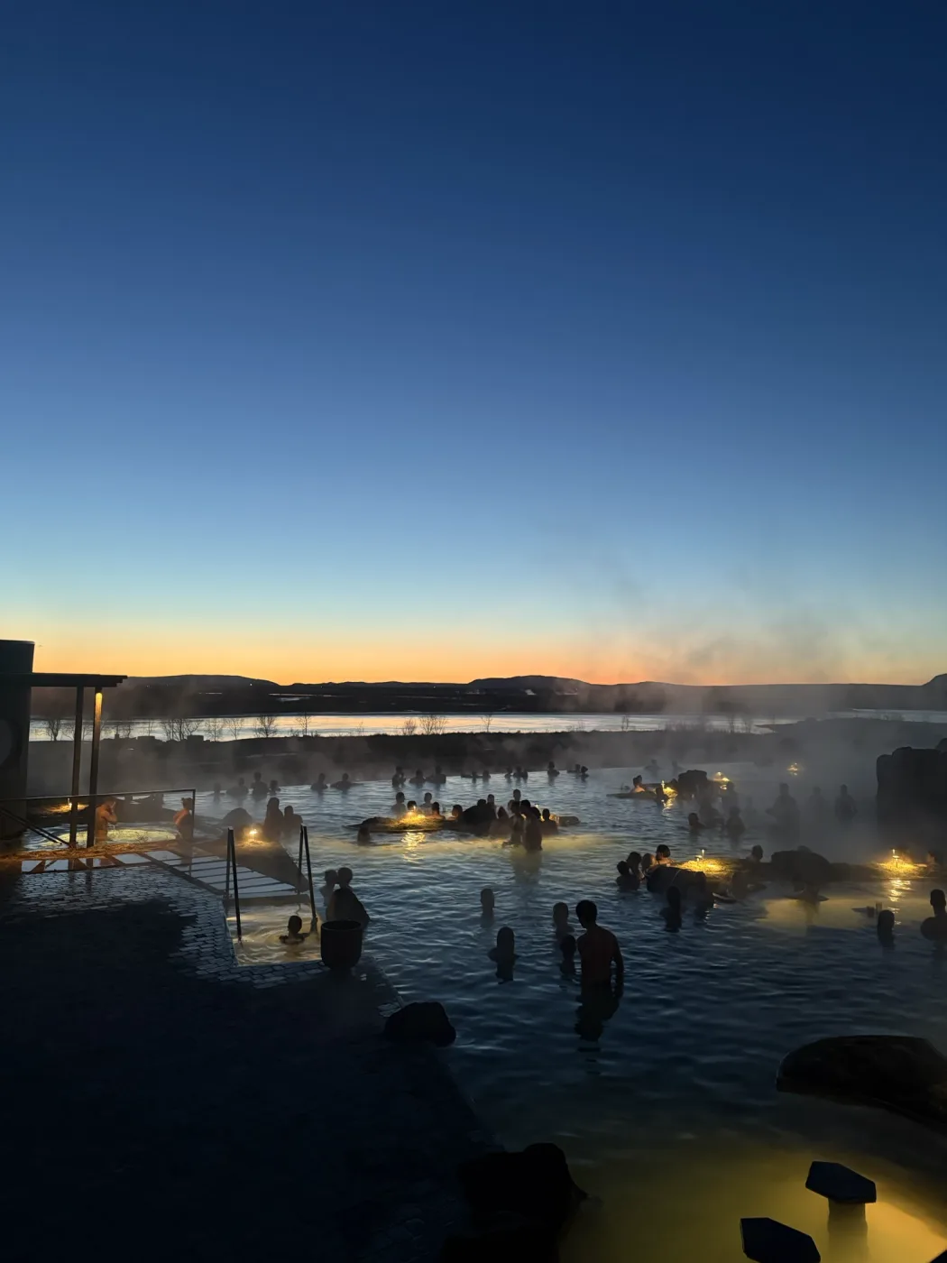 Bathers in a steaming geothermal lagoon at dusk, amber light on the horizon